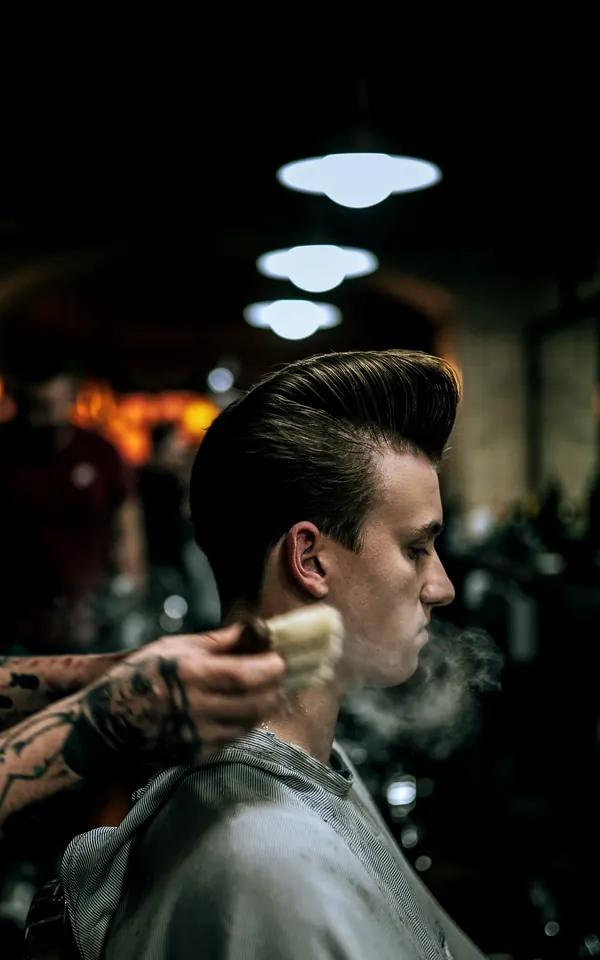 A barber finishing a pompadour with a neck brush under low pendant lights